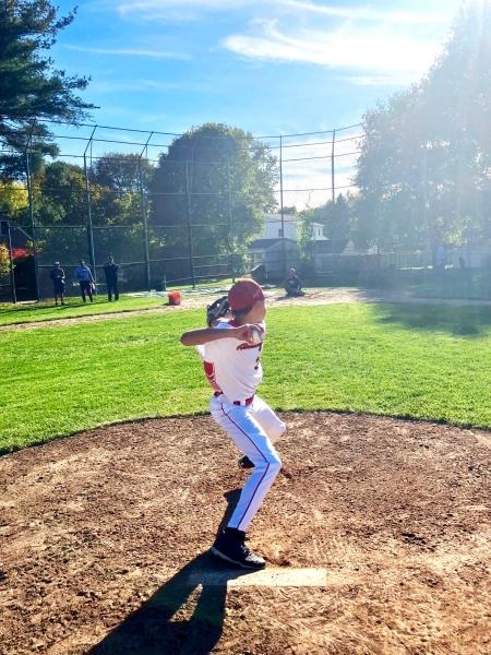 An RBRL pitcher in red and white winds up on the mound at Pete Moscariello Ballpark, the late-day sun behind him.